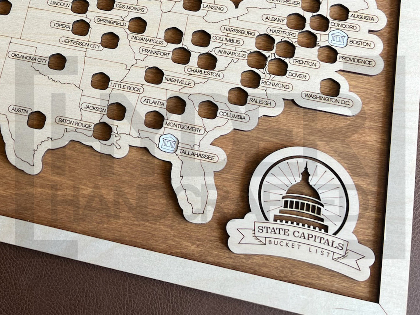 A framed State Capitals bucket list map with a natural wood finish, white tokens, and a sawtooth hanger. The map shows a list of state capitals and has a seal stamp with the text 'STATE CAPITALS' on it.