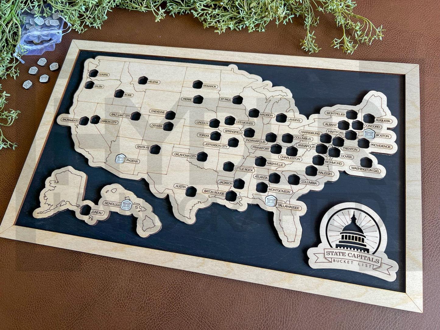 A framed State Capitals bucket list map with a natural wood finish, white tokens, and black stain background. The map shows a list of state capitals and has a seal stamp with the text 'STATE CAPITALS' on it.