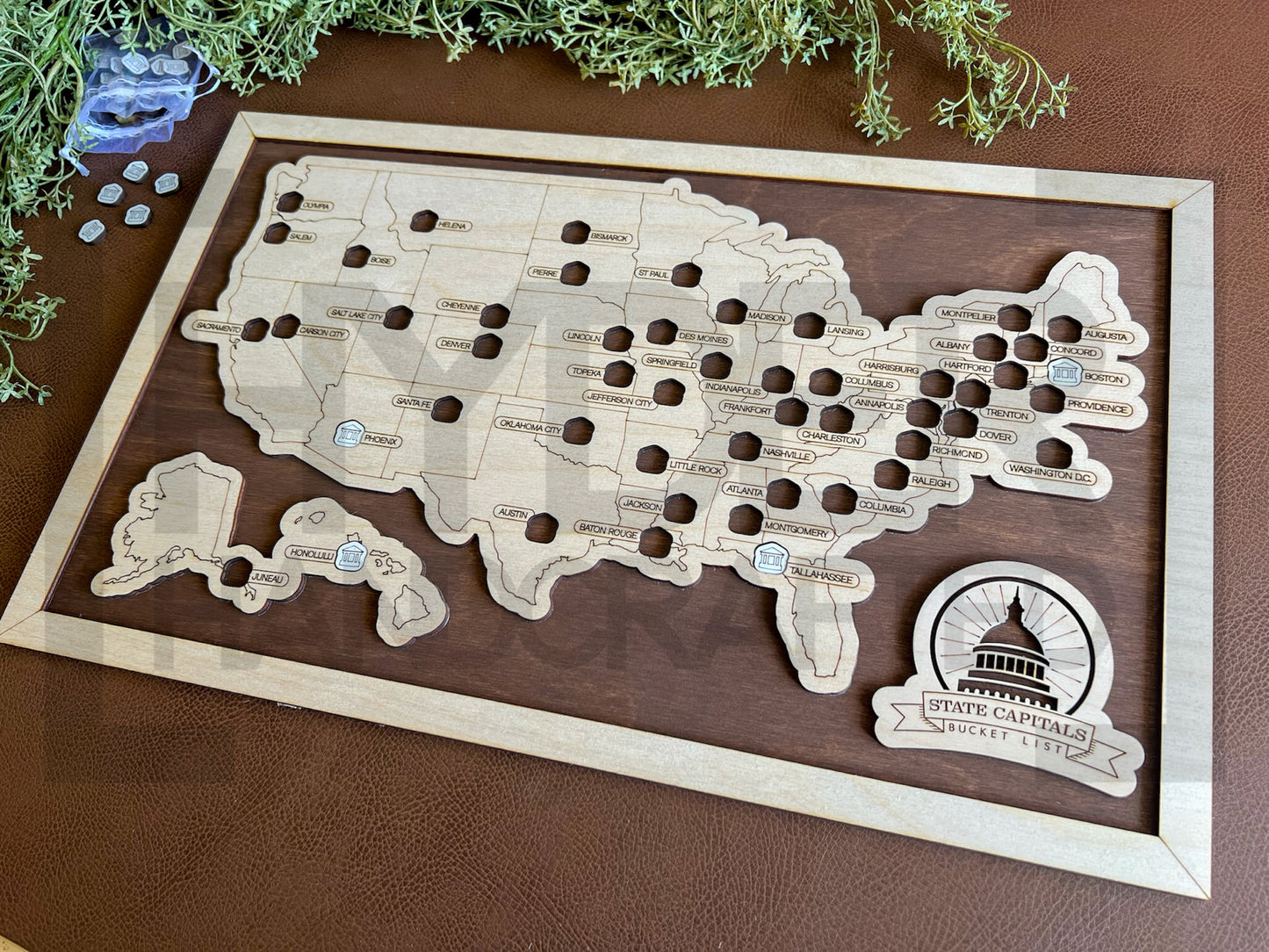 A framed State Capitals bucket list map with a natural wood finish, white tokens, and dark wood stain backing. The map shows a list of state capitals and has a seal stamp with the text 'STATE CAPITALS' on it.