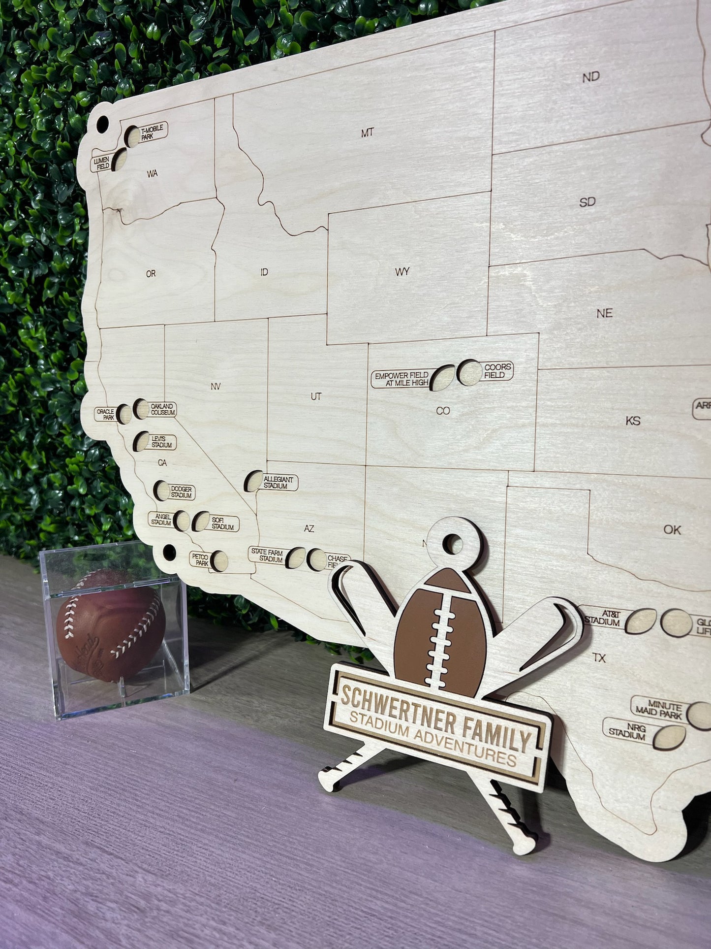 Handcrafted wooden multi-sport map with a football and baseball decoration, displayed on a stand against a greenery backdrop.