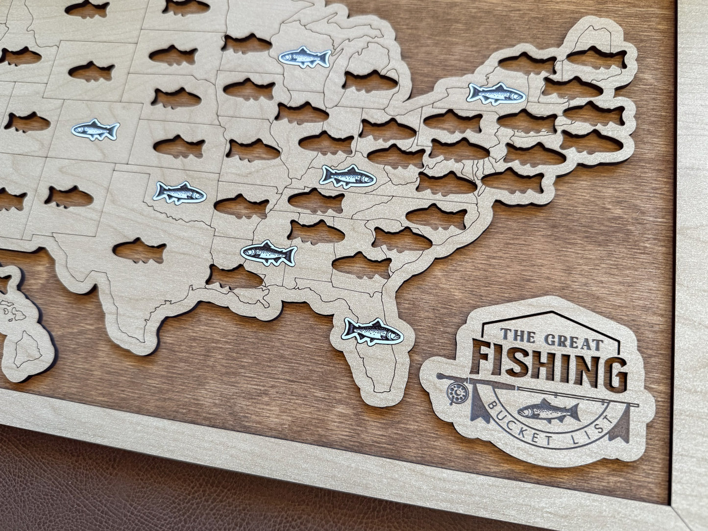 Wooden map of the United States with cut-out fish shapes and 'The Great Fishing Bucket List' logo.