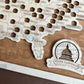 A framed State Capitals bucket list map with a natural wood finish, white tokens, and a sawtooth hanger. The map shows a list of state capitals and has a seal stamp with the text 'STATE CAPITALS' on it.