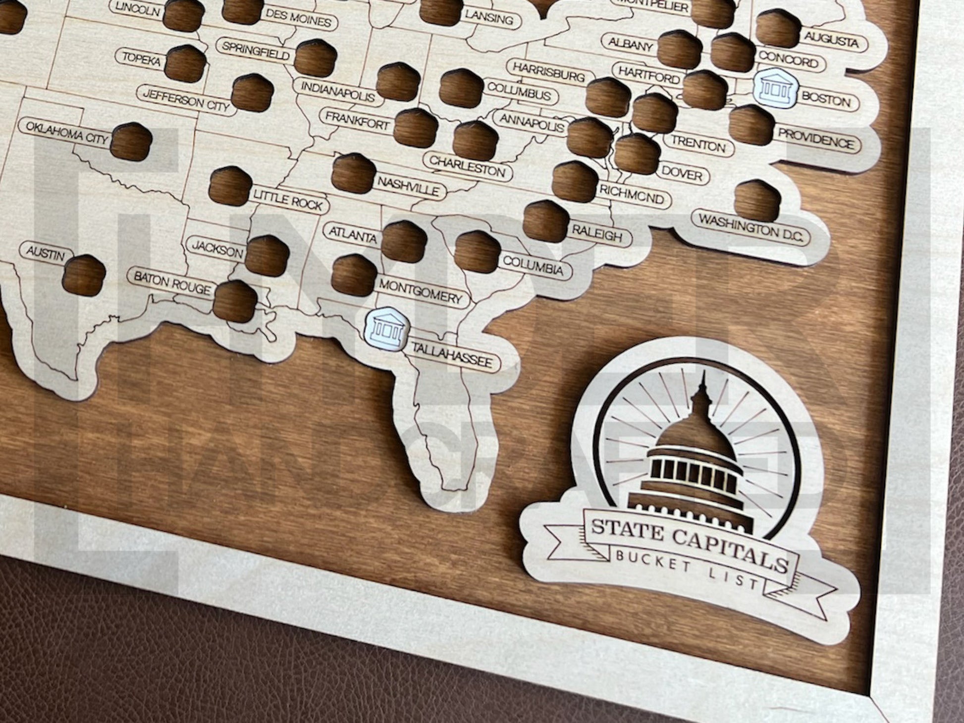 A framed State Capitals bucket list map with a natural wood finish, white tokens, and a sawtooth hanger. The map shows a list of state capitals and has a seal stamp with the text 'STATE CAPITALS' on it.
