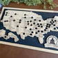 A framed State Capitals bucket list map with a natural wood finish, white tokens, and black stain background. The map shows a list of state capitals and has a seal stamp with the text 'STATE CAPITALS' on it.