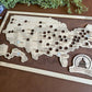 A framed State Capitals bucket list map with a natural wood finish, white tokens, and dark wood stain backing. The map shows a list of state capitals and has a seal stamp with the text 'STATE CAPITALS' on it.