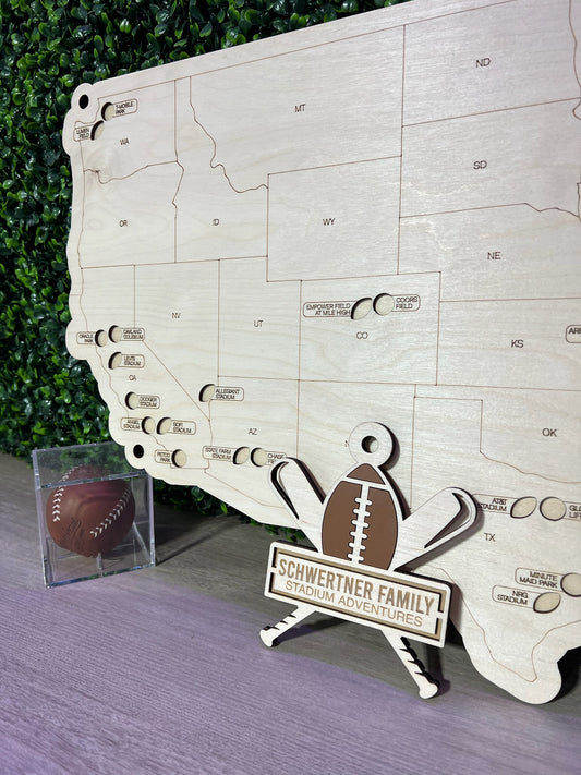 Handcrafted wooden multi-sport map with a football and baseball decoration, displayed on a stand against a greenery backdrop.