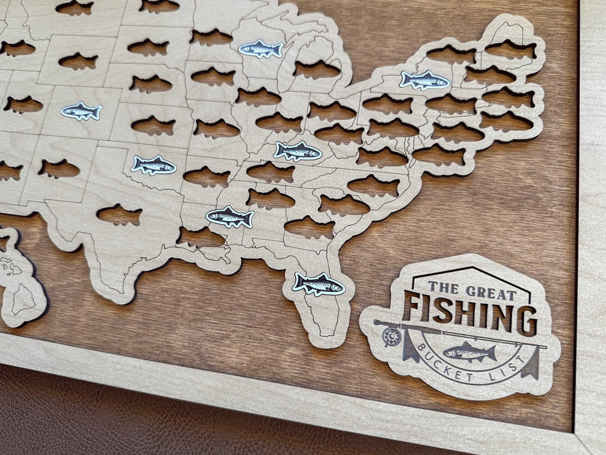Wooden map of the United States with cut-out fish shapes and 'The Great Fishing Bucket List' logo.