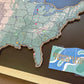 Map of the United States with pins and a personalized travel map card on a wooden surface.