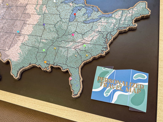 Map of the United States with pins and a personalized travel map card on a wooden surface.