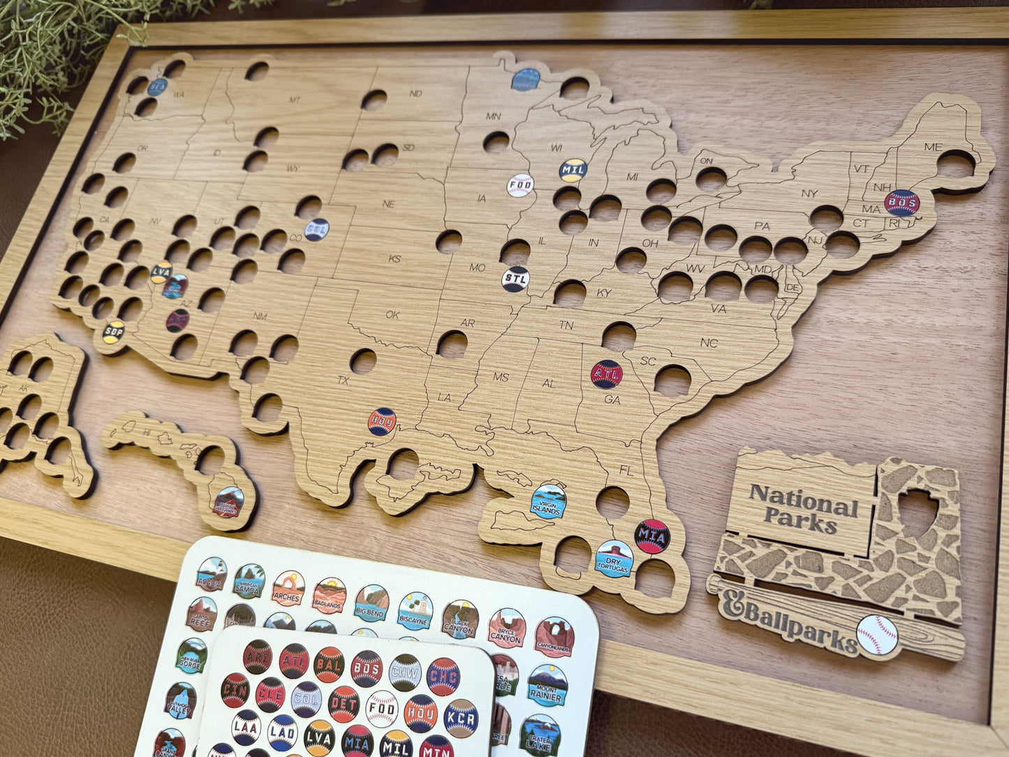 Wooden map of the United States with tracking token stickers and a small sign labeled 'National Parks' and baseball stadiums on a wooden surface.