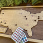 Wooden map of the United States with pins and a plank of sticker tokens on a wooden surface.